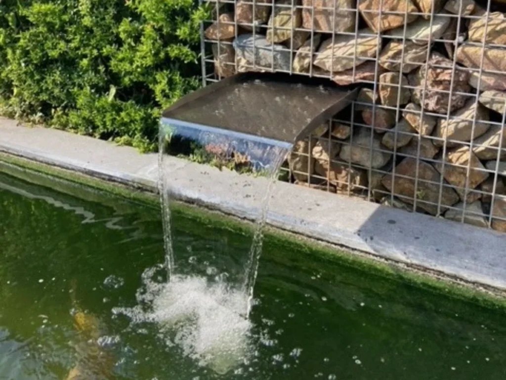 Edelstahl-Bachlauf-Wasserfall, eingebaut in eine Gabionenwand, mit sanftem Wasserfluss in Gartenteich