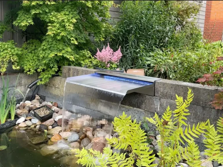 Edelstahl-Wasserfall mit LED-Beleuchtung an einer Mauer über einem Teich mit Naturbepflanzung
