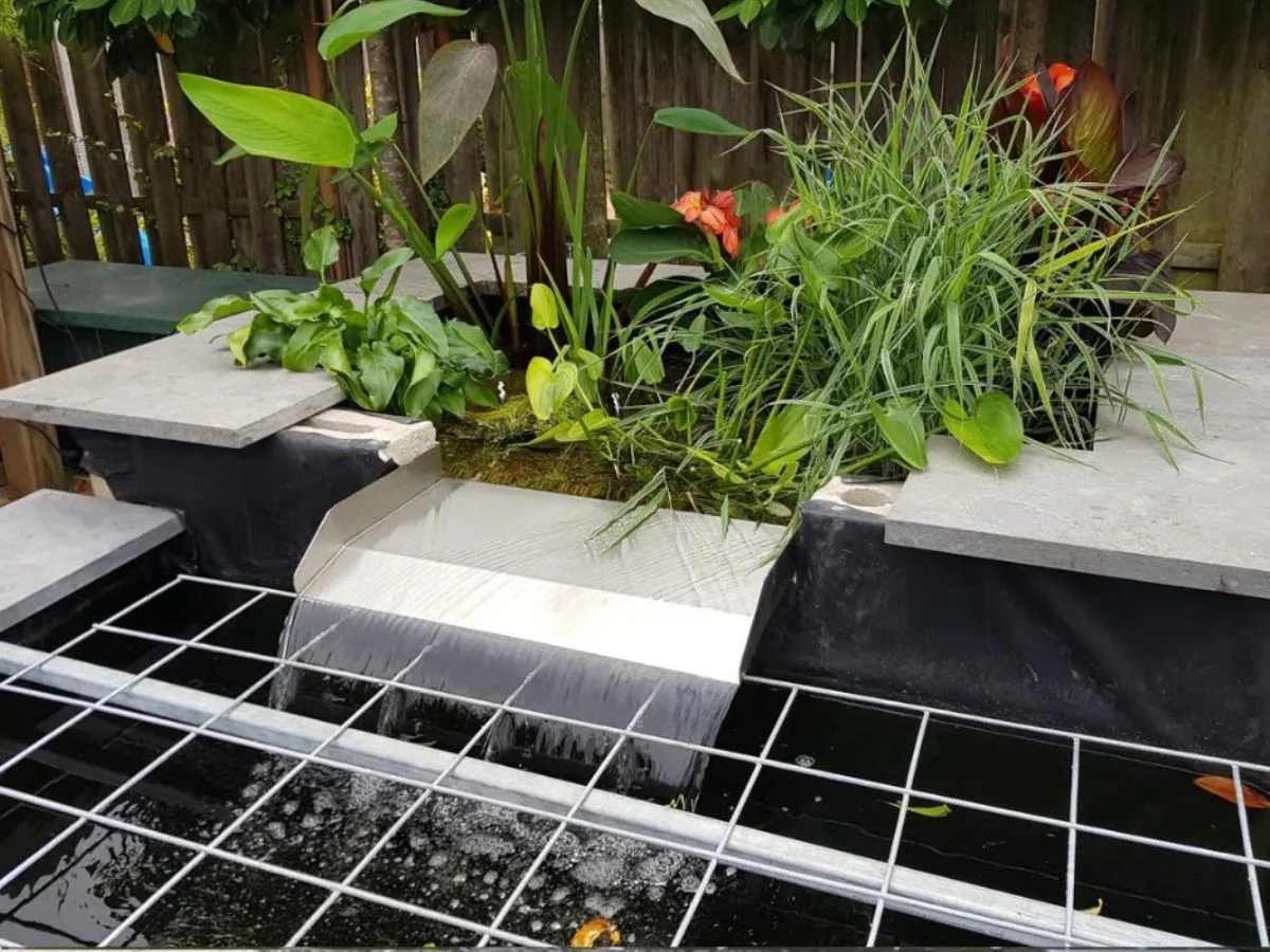 Moderner Edelstahl Überlaufwasserfall, eingebaut zwischen Natursteinplatten, mit üppiger Bepflanzung am Gartenteich.