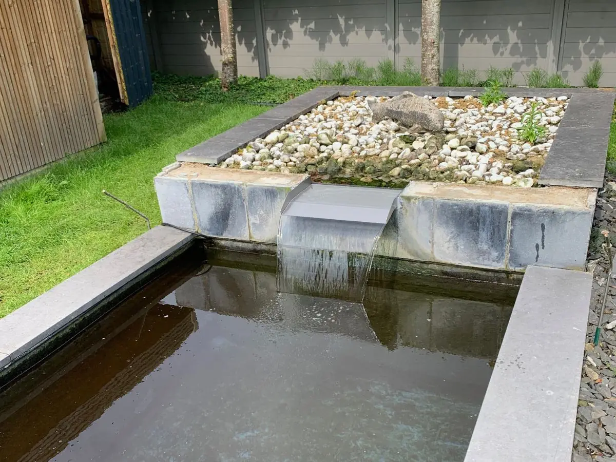 Edelstahl Überlaufwasserfall in Gartenteichanlage mit Kies, Steinumrandung und dekorativem Fischornament.
