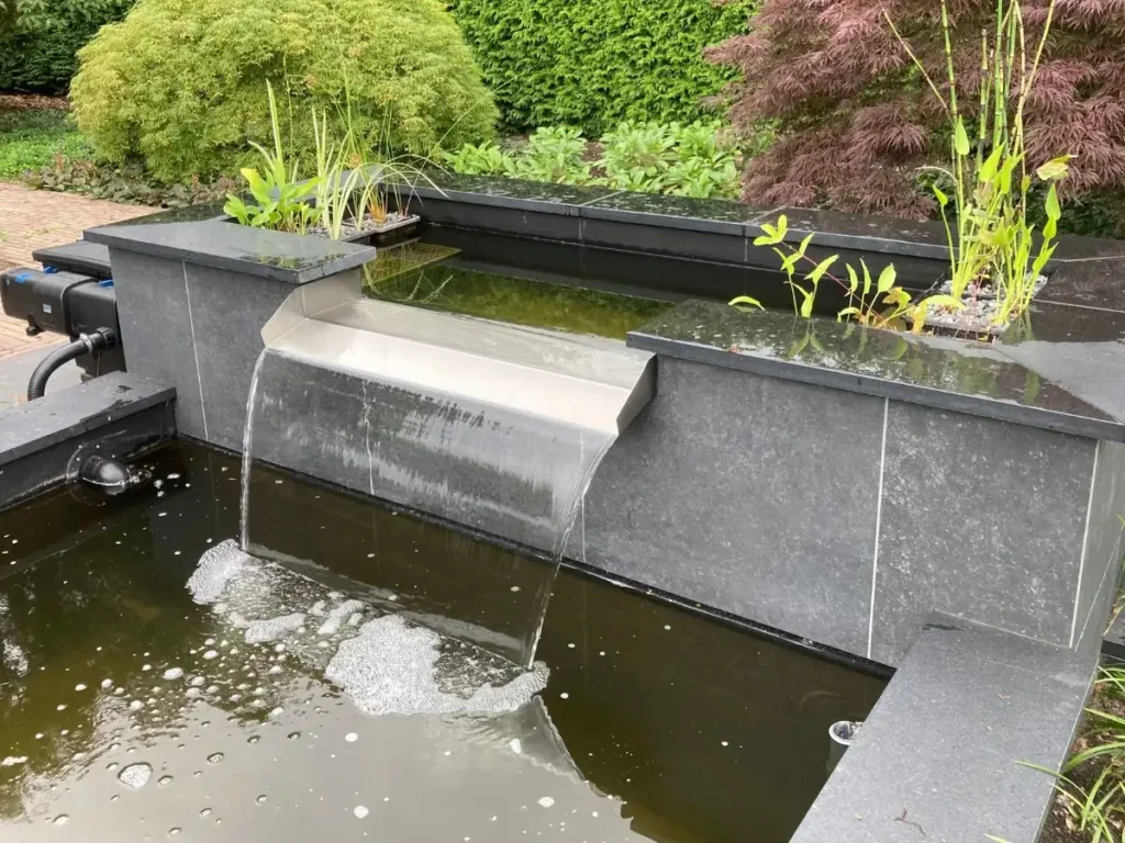 Edelstahl Überlaufwasserfall mit gleichmäßigem Wasserfluss in einen modernen Gartenteich mit dunkler Steinumrandung.