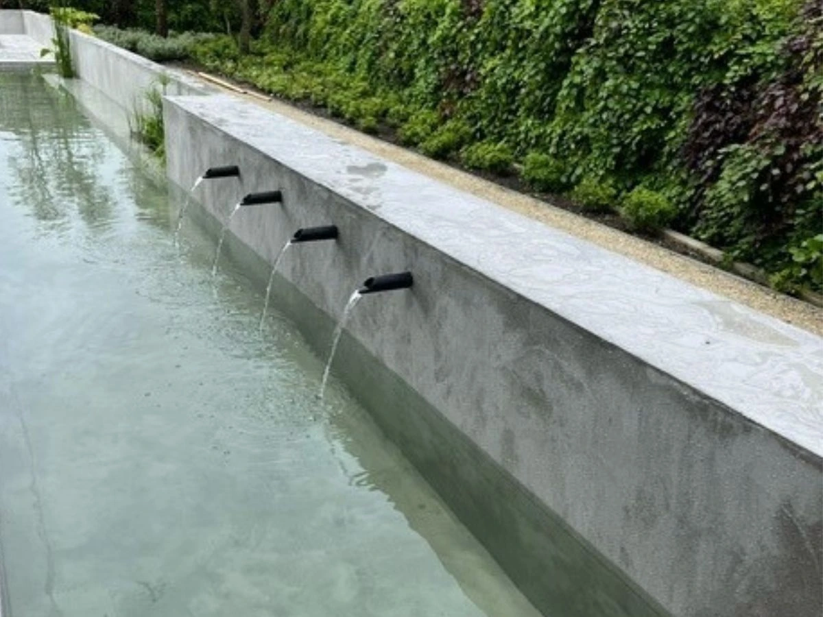 Vier gerade Edelstahl-Wasserausläufe in moderner Betonmauer mit Wasserzufuhr in flachem Gartenteich